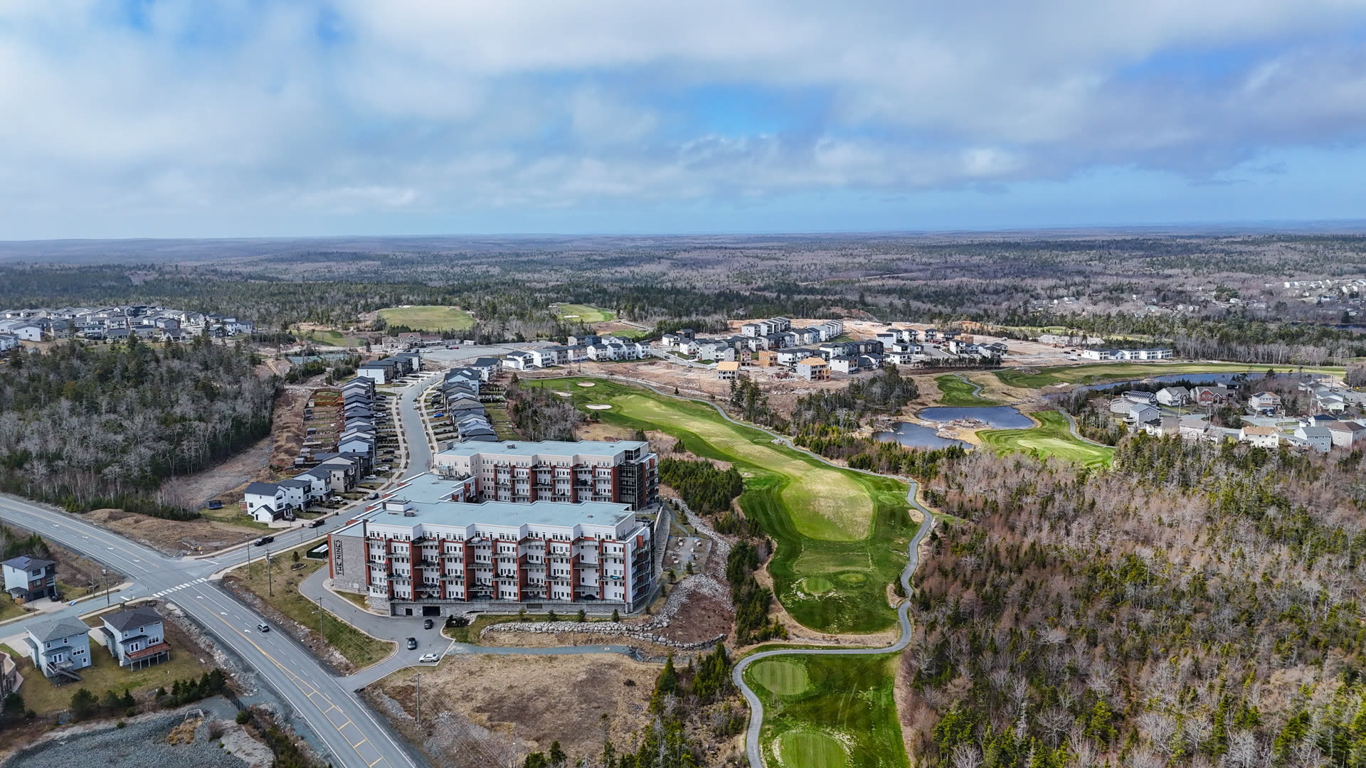 Aerial view of a modern mid-rise residence beside green golf fairways and surrounding forest.